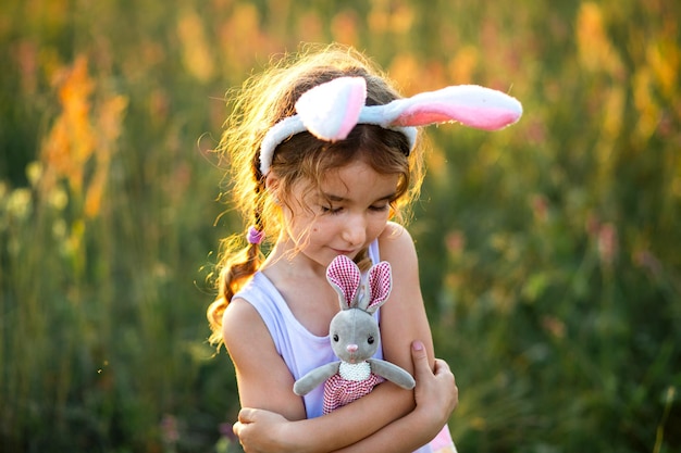 Cute 5-year-old girl with rabbit ears gently hugs a toy rabbit in nature in a blooming field in summer with golden sunlight. Easter, Easter bunny, childhood, happy child, springtime.