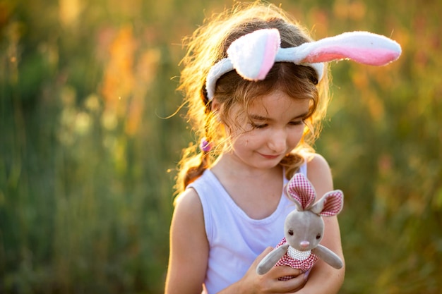 Cute 5-year-old girl with rabbit ears gently hugs a toy rabbit in nature in a blooming field in summer with golden sunlight. Easter, Easter bunny, childhood, happy child, springtime.