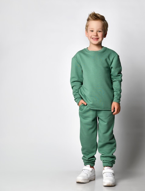 Cute active little boy in trendy sports green clothes standing with hand in pocket showing emotions of happiness and comfort Positive cheerful childish expression