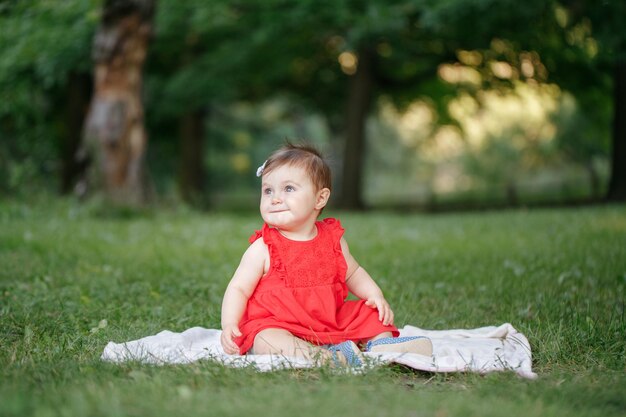 Cute adorable baby girl in red dress sitting on white blanket in park outdoors funny child toddler