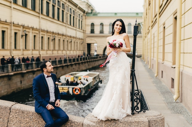 Cute adorable bride with long dark hair, wears wedding dress, holds beautiful bouquete of flowers, stands on bridge and her husband who sits near her