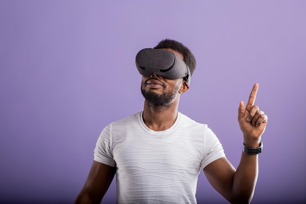 Cute african american man in VR glasses, playing video games with virtual reality headset, interacting with invisible screen on lilac background with copy space. 3D technology, VR concept.