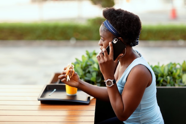 Cute African woman having lunch outdoors in city cafe. A girl is talking on the phone during lunch.