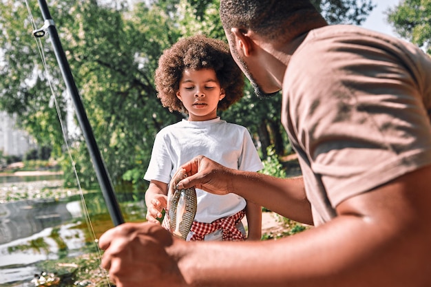 Cute AfroAmerican child looking st the fish in hands of his dad on a fishing trip