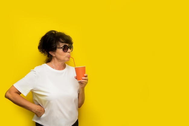 Cute aged woman in sunglasses drinking from a straw a cocktail from a paper glass on a yellow background with copy space