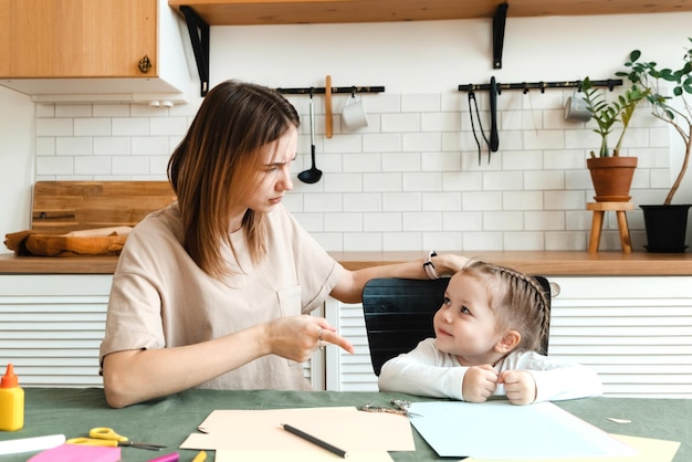 Cute angry little preschool girl doing creative homework with her mom while sitting at the table in the living room and arguing Problems in making crafts for kindergarten