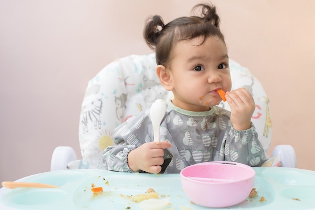 A cute Asian baby girl sitting on dining table practice eat carrot and another food by her self, Baby-Led Weaning concept