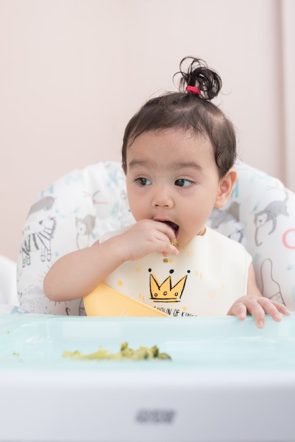 A Cute Asian baby girl wearing  baby apron sitting on dining table eat broccoli by her self, Baby Led Weaning concept