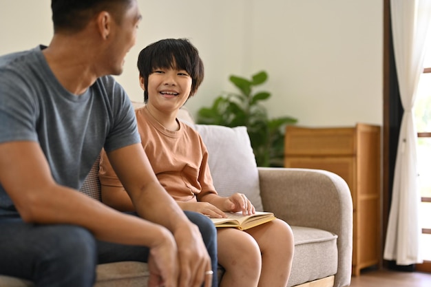 Cute Asian boy sits on a sofa with his dad in the living room spending family time together