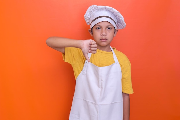 Cute Asian boy wearing chef uniform having sad expression while showing thumb down