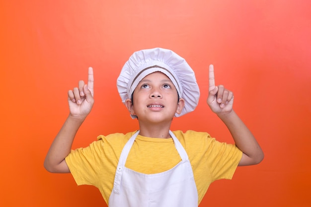 Cute Asian boy wearing chef uniform pointing finger up and  arms raised with eyes looking up