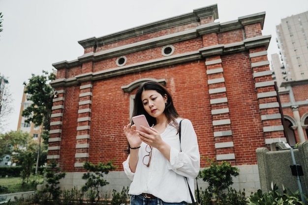 Cute asian chinese girl traveler in casual wear standing in front of red brick wall background on sunny day using mobile phone searching way online. lady traveler reading map internet find direction