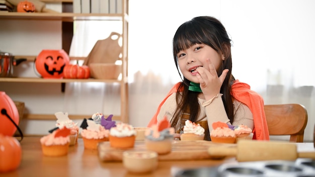 Cute Asian girl in Halloween fancy costume tasting a Halloween cupcake in the kitchen