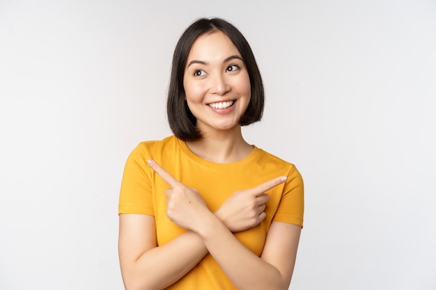 Cute asian girl pointing fingers sideways showing left and right promo two choices variants of products standing in yellow tshirt over white background