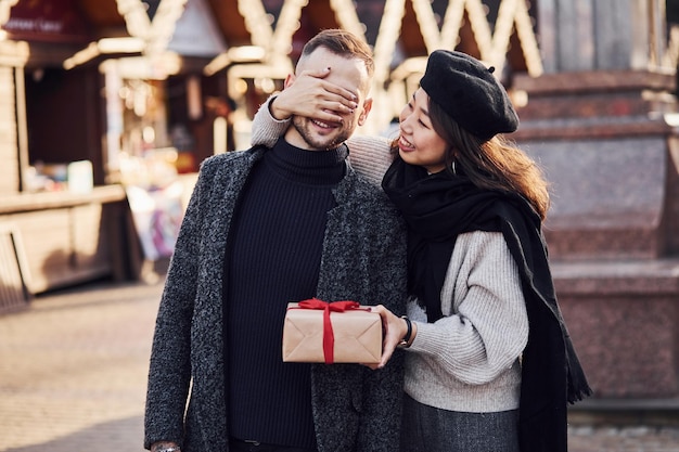 Cute asian girlfriend making surprise and giving gift box to her caucasian man.