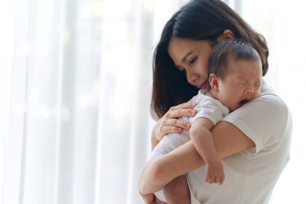 Cute Asian infant baby on mothers hands standing in the room near the window. 