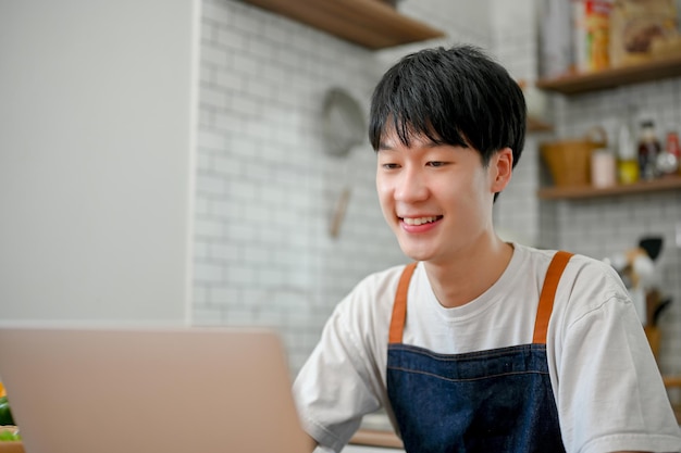 Cute Asian man in apron using laptop in his kitchen room searching food recipe on the internet