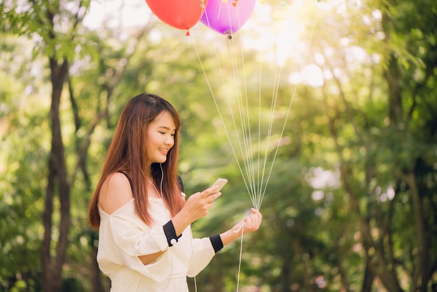 Cute asian woman is reading pleasant text message on mobile phone while sitting in park in warm spring day,gorgeous female listening to music in headphones and searching information on cell telephone.