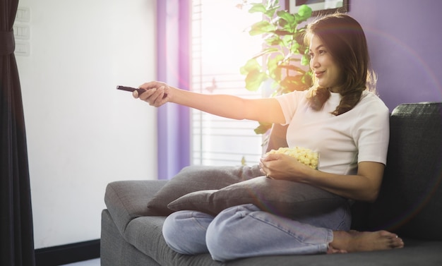 Cute Asian woman sitting on sofa with relax and cozy gesture holding remote control aiming to tv, watching television and smile with fun and happy in living room with sunlight and light flare.