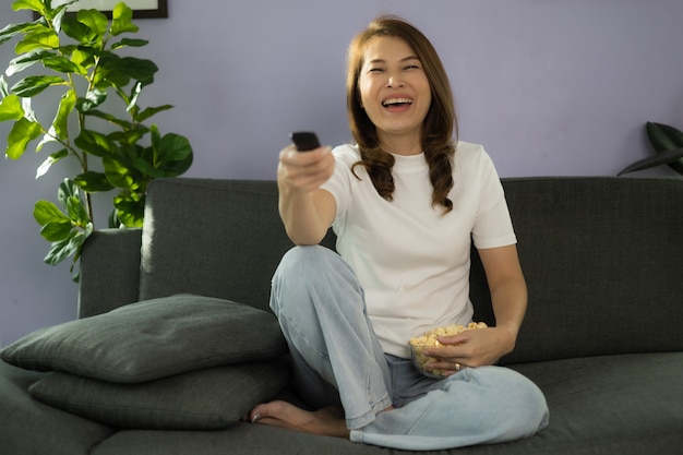 Cute Asian woman sitting on sofa with relax and cozy gesture holding remote control, watching television and laugh with fun and happy in home living room with sunlight from beside of her body.