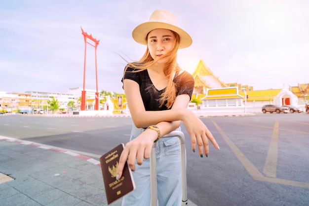 A cute Asian woman stands in the middle of Bangkok's tourist attractions in Thailand With a suitcase and passport The background is Giant Swing