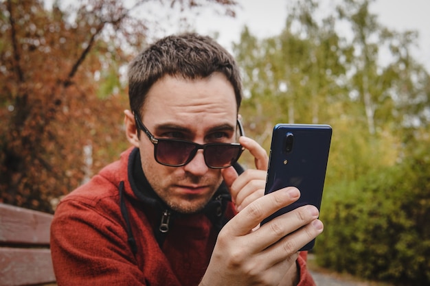 Cute attractive handsome serious guy wearing casual clothes and sunglasses outside holding smartphone reading sms. Work through smartphone. Correspondence in social network. Student in Park chatting.