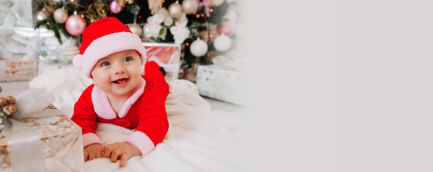 A cute baby 6 months old in a Santa costume lies on a blanket near the Christmas tree christmas 2022