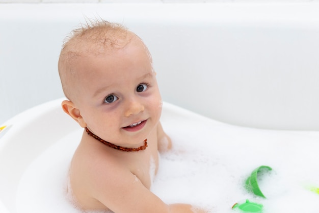 A cute baby boy is washing in the bathtub, playing with green plastic cup toy in foam bubbles.