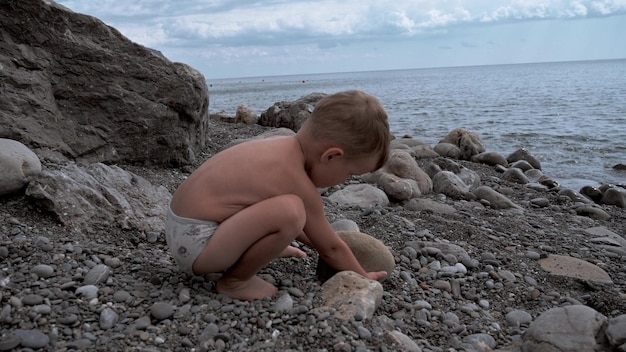 Cute baby boy playing on the seashore with rocks and pebbles The kid is having a good time on a family vacation Slow motion video