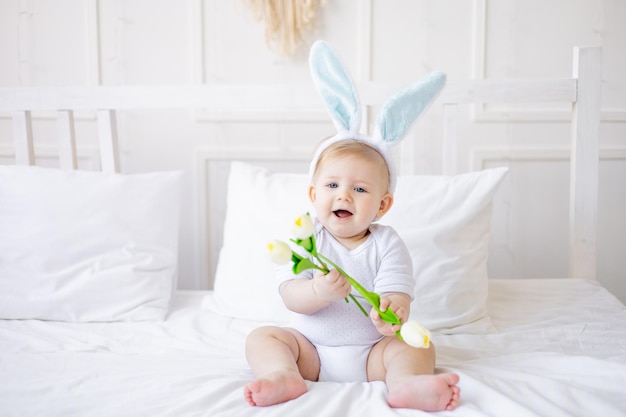 Cute baby boy with bunny ears and tulip flowers on a white bed at home little blonde baby holding flowers happy easter concept