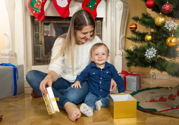 Cute baby boy with his mother opening gift boxes under Christmas tree at living room