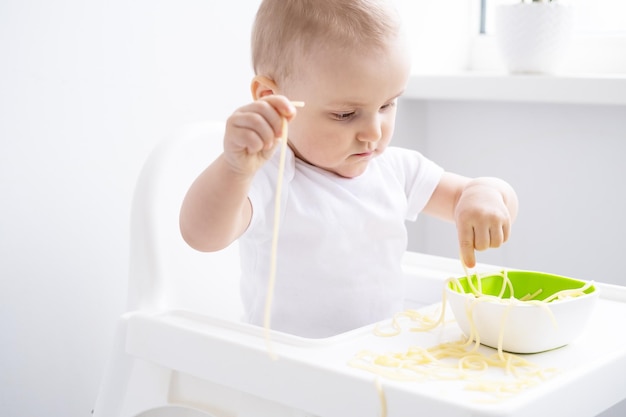 cute baby girl eating spaghetti pasta sitting in baby chair on white kitchen