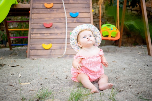 Cute baby girl sitting on the sand on the playground in summer in a panama hat and bare feet