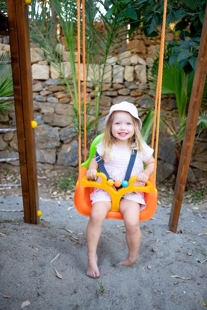 Cute baby girl swings on a swing and laughs in the summer in a panama hat