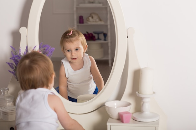 Cute baby girl watching her reflection in a white bedroom with a round mirror