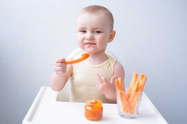 Cute baby girl in yellow bodysuit sitting in childs chair eating carrot slices and vegetables purees