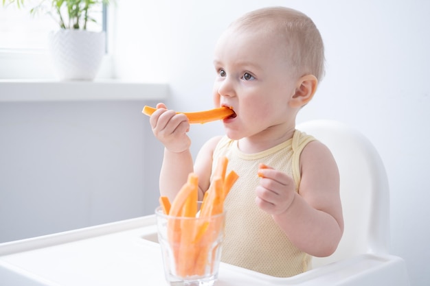 cute baby girl in yellow bodysuit sitting in childs chair eating carrot slices