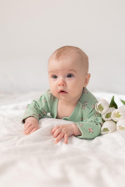 Cute baby in a green cotton bodysuit at home on a white bed with tulips. Spring, Mother's Day, March