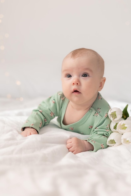 Cute baby in a green cotton bodysuit at home on a white bed with tulips. Spring, Mother's Day, March