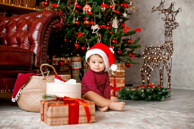 Cute baby Santa sits at home near the Christmas tree with gifts