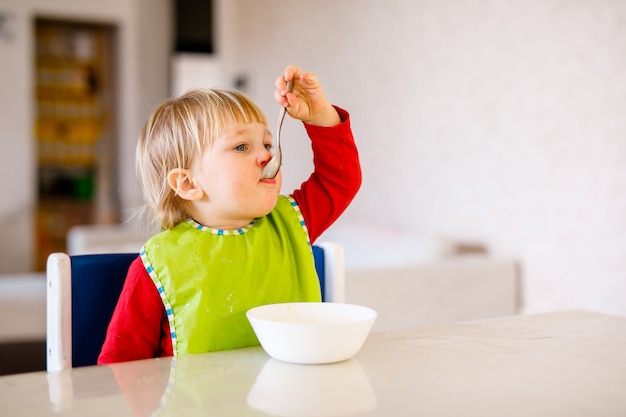 Cute baby sitting on high children chair and eating vegetable alone in white kitchen.