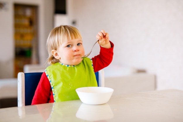 Cute baby sitting on high children chair and eating vegetable alone in white kitchen.