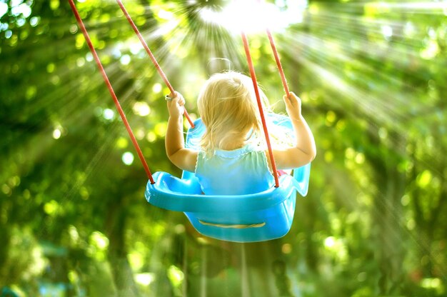 cute baby toddler swinging in the park with high trees and sun rays coming through