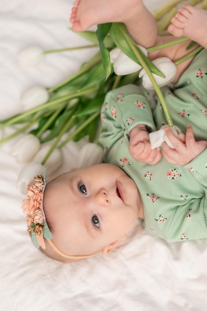 Cute baby with a floral headband and a green cotton bodysuit at home on a white bed with tulips. Spr