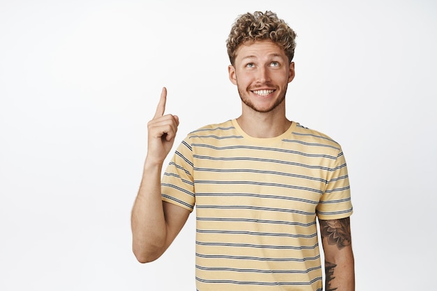 Cute bearded guy with blond hair smiling happy while looking and pointing up picking product from advertisement white background