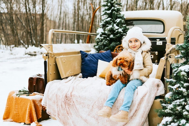 Cute beautiful little Armenian girl in winter coat and knitted hat and pet poodle dog sit near beige retro pickup truck decorated for Christmas and New Year vintage interior suitcases skis sledge