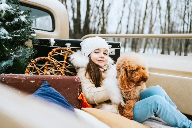 Cute beautiful little Armenian girl in winter coat and knitted hat and pet poodle dog sit near beige retro pickup truck decorated for Christmas and New Year vintage interior suitcases skis sledge