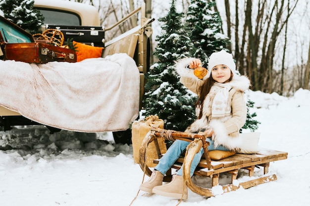 Cute beautiful little Armenian girl in winter coat and knitted hat sit near beige retro pickup truck decorated Christmas and New Year vintage interior items suitcases skis sledge ornaments travel