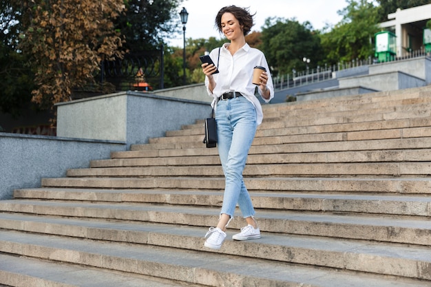 Cute beautiful woman walking on the street using mobile phone holding coffee