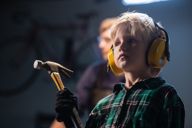 Cute blond child boy with a hammer in hand in a carpenter's workshop, close-up portrait.